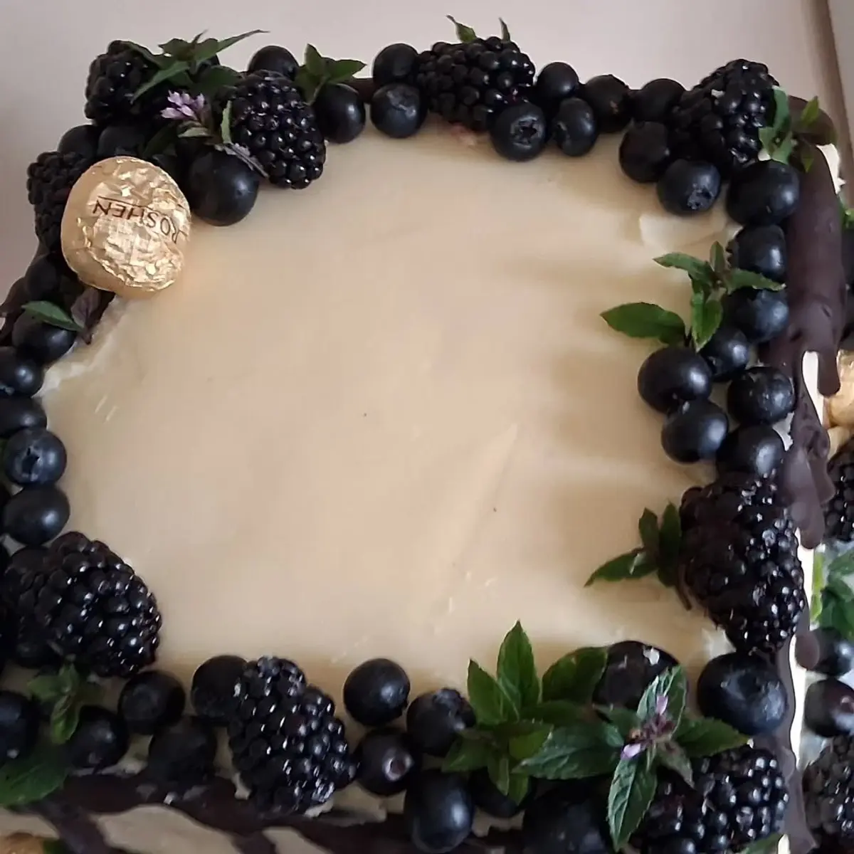 Top-down view of a square cream-iced cake bordered with blackberries, blueberries, mint and a chocolate drip