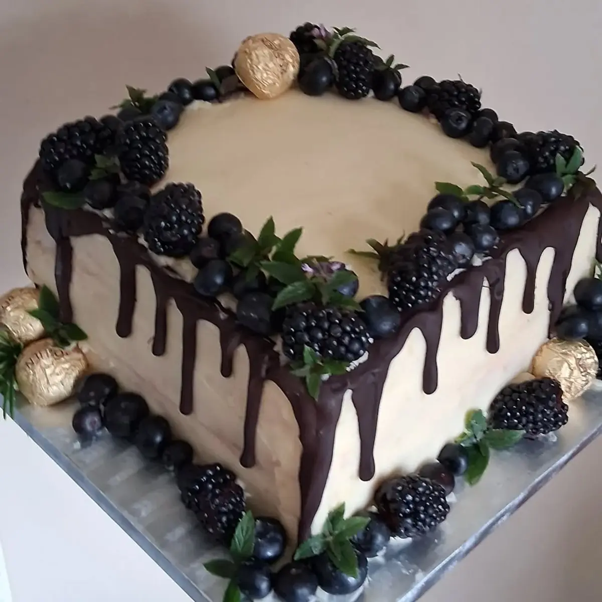 A bespoke celebration cake with cream icing, a dark chocolate drip, fresh blackberries and blueberries, mint and gold-wrapped chocolates