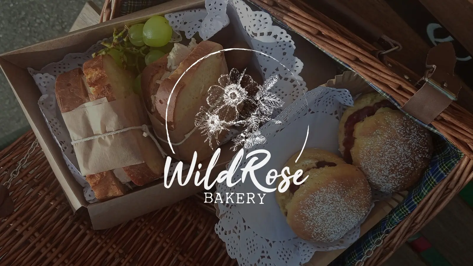 A Wild Rose Bakery hamper in a wicker basket - sliced breads, filled buns and scones, and grapes on white doilies